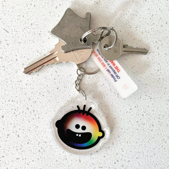 Our ‘Love for all’ charm featuring a rainbow Cheeky Pebble face on a silver keychain and ring. Displayed on a quartz background..